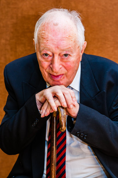 照片中，一位身穿深色西装、白衬衫、系着蓝红色条纹领带的白人老人坐在椅子上，双手交叉抱着头，拄着一根拐杖，背景是亮棕色图片下载