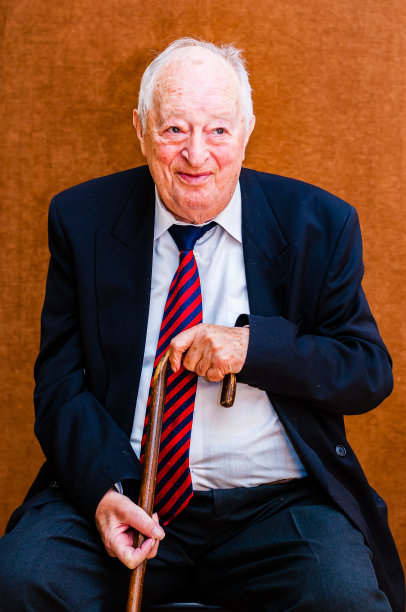 照片中的白人老人身穿深色西装、白色衬衫，系着蓝红色条纹领带，面带微笑地坐在椅子上，手里拿着一根拐杖，背景是亮棕色图片下载