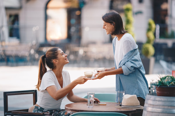 一个女人在室外给另一个女人递热饮。他们彼此很友好。桌子上有一部智能手机和一顶草帽。图片下载