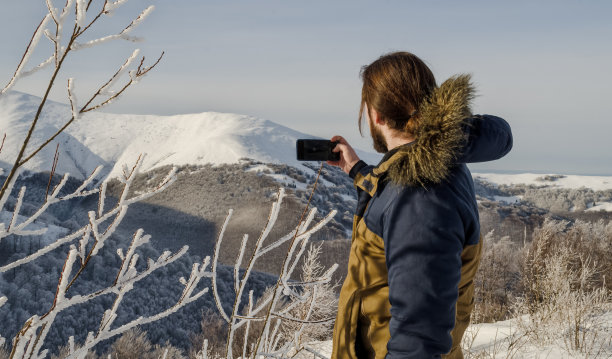 一个穿着冬季夹克的男人，在群山的背景下，手里拿着手机图片下载