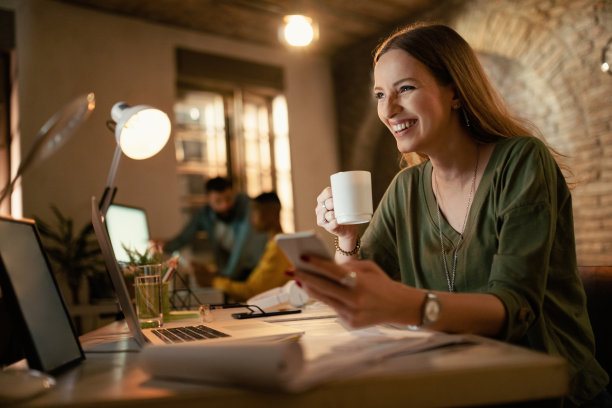 快乐的女商人一边喝咖啡一边在办公室用手机。图片下载