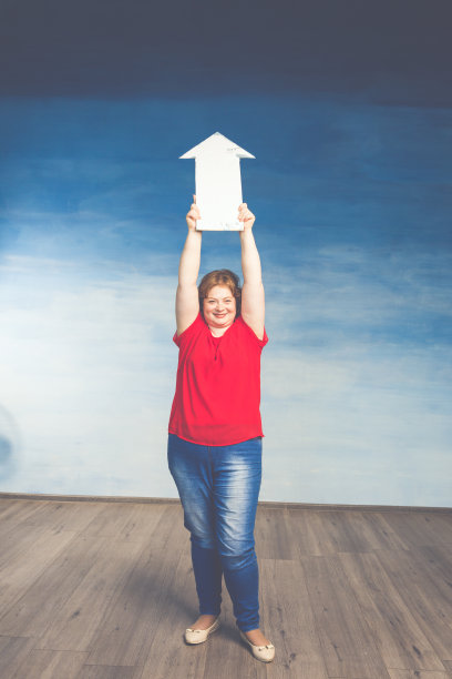 穿着红色t恤和牛仔裤的漂亮红发胖女人举着一个向上的箭头图片下载