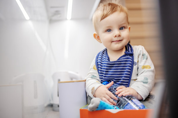 可爱的小男孩，有着美丽的大蓝眼睛，坐在厨房柜台上的盒子里，拿着一瓶水，看着相机。图片下载