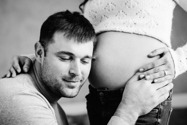 英俊的男人听着他美丽的怀孕妻子的肚子微笑着。女人和有爱心的男人在家里抱着肚子。爱的夫妻。亲子关系的概念。婴儿洗澡。黑白照片。图片下载