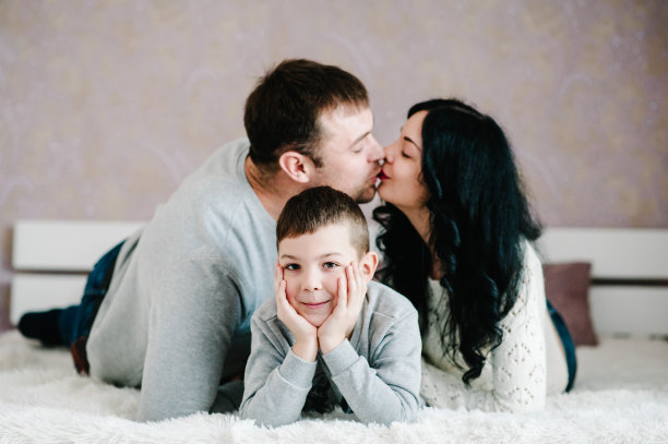 幸福的家庭照片。丈夫，妻子和儿子在家里拥抱。快乐的儿子看着妈妈亲吻爸爸，一起享受他们的节日时光。图片下载