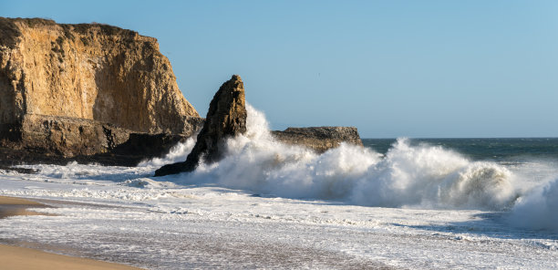 在蓝天的沙滩上，强烈的海浪撞击岩石形成的景象图片下载