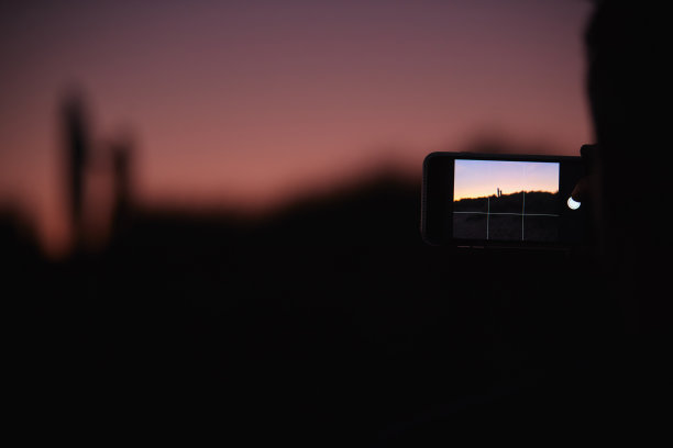在手机上拍摄剪影冲浪者站在沙丘上和在日落时持有冲浪板的照片图片下载