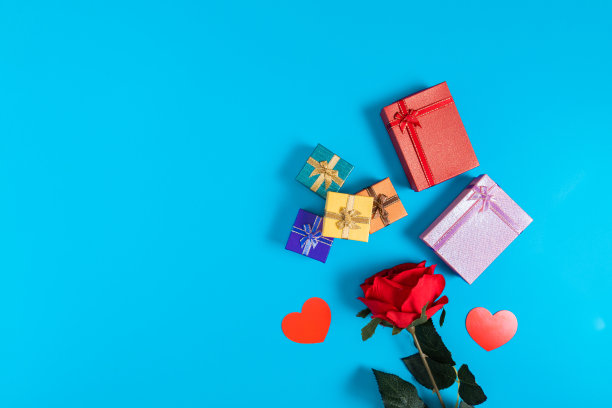 6个礼品盒，分别是红色、粉色、深蓝色、橙色、黄色和绿色，放在蓝色背景上，位于图片的中心。在图片的底部有红玫瑰和心形，这是情人节图片下载