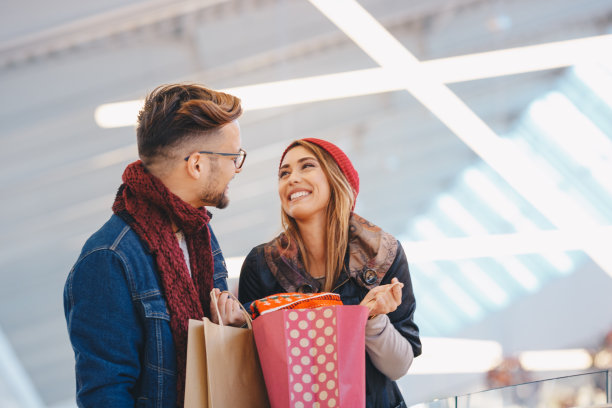 一位年轻女子向支持她的丈夫展示她买的东西。丈夫在对她微笑。这个场景是在购物中心拍摄的。素材图片
