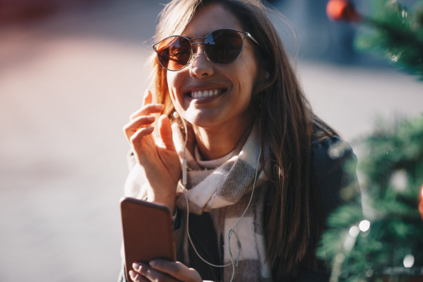 圣诞节期间在户外听音乐的女人。她闭上眼睛，戴着耳机，拿着智能手机。图片下载