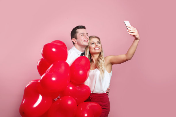 一对美丽的年轻夫妇用手机自拍，粉色背景上站着红色气球图片下载