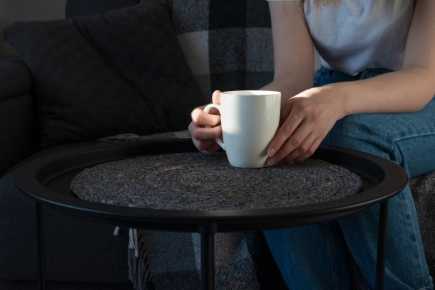 在阳光明媚的日子里，在室内沙发上的咖啡桌上，女人手拿着白色的杯子。在家休息，生活方式。图片下载