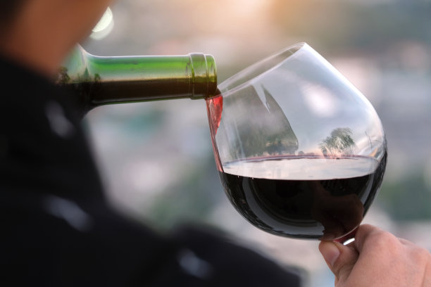 在一个浪漫的餐厅里，年轻人的手把红酒倒进了一杯红酒里。夕阳和现代城市背景图片下载