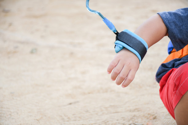 单身男孩在户外活动或玩耍时用蓝色皮带牵着他的手，背景是沙滩。儿童皮带安全概念。图片下载