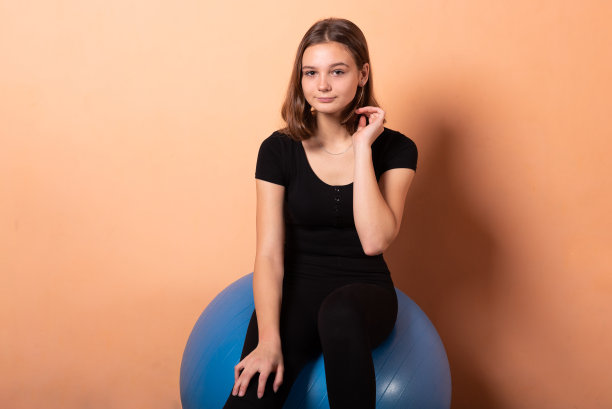 女孩坐在健身球上，背景是浅橙色。图片下载