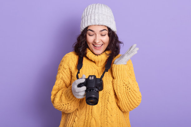 迷人的女孩看着照片在相机，摆姿势在淡紫色的背景，女性穿着温暖的针织黄色毛衣，手套和帽子，使照片，看起来快乐，保持张嘴。爱好的概念。图片下载