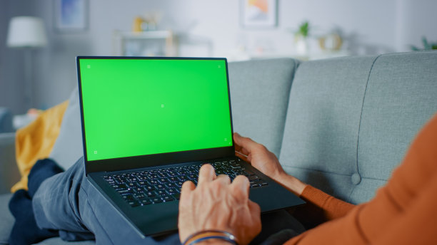 一名男子躺在家里的沙发上，使用带有绿色模拟屏幕的笔记本电脑。客厅里的无名氏的肩膀照片。图片下载