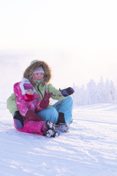 3岁的女孩和孩子穿着冬季运动服坐在雪山顶上。妈妈把手伸到远处。图片下载