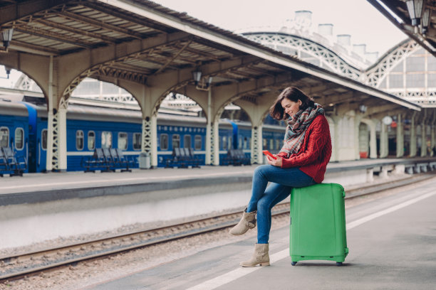 照片中，一名年轻的欧洲女子穿着休闲服装，坐在行李旁，拿着手机，开心地接收短信，在站台摆姿势，享受空闲时间，等待火车。旅游的概念图片下载