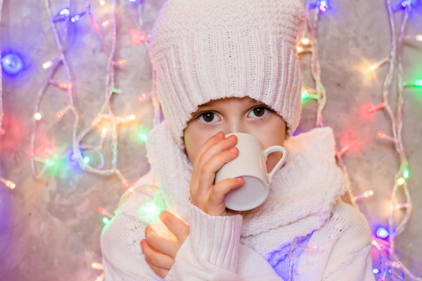 穿着白色针织毛衣戴着帽子的冻小女孩在一个彩色花环的背景下喝着一杯热腾腾的饮料。冷和暖的概念图片下载