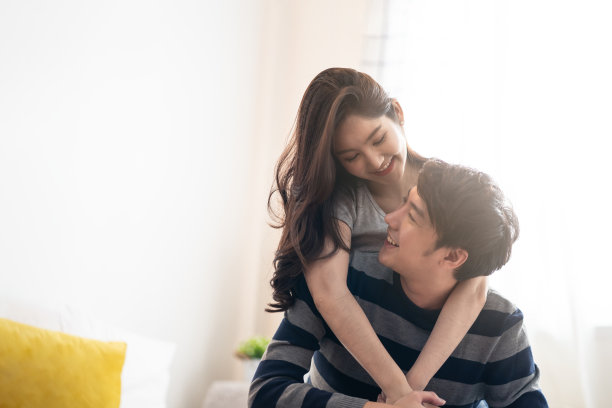 亚洲年轻夫妇，男人和女人一起待在卧室里。女孩从后面拥抱着男孩，互相看着对方感觉爱的幸福，面带微笑。年轻女孩和男孩概念的关系。图片下载