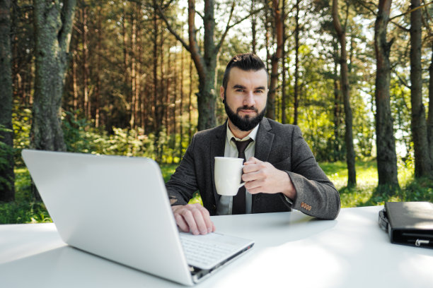 年轻有吸引力的商人西装革履坐在办公桌前，在电脑上工作。用白色的杯子喝咖啡。以绿树、自然和公园为背景。图片下载