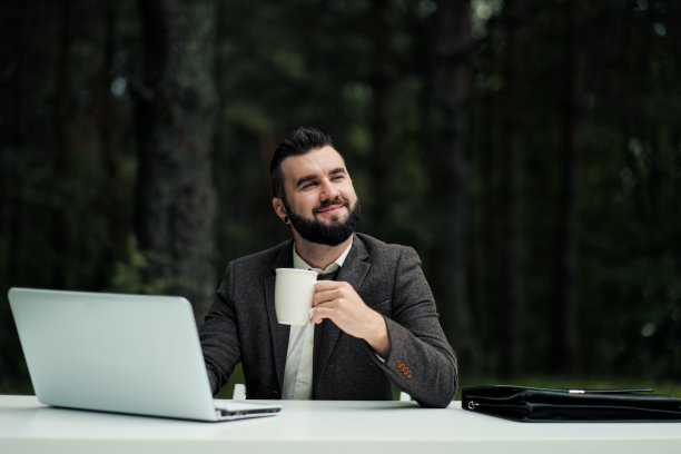 年轻有吸引力的商人西装革履坐在办公桌前，在电脑上工作。用白色的杯子喝咖啡。以绿树、自然和公园为背景。图片下载