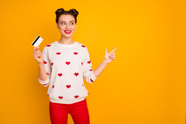 有趣感兴趣的女士的照片拿着塑料信用卡指示手指空白推荐快速支付方式穿心形套头衫红色裤子孤立的黄色背景图片下载