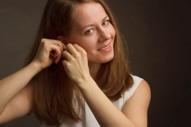 画室里一个穿着白色连衣裙的金发模特的肖像，背景是黑色的。扣上耳环，手放在耳朵上图片下载