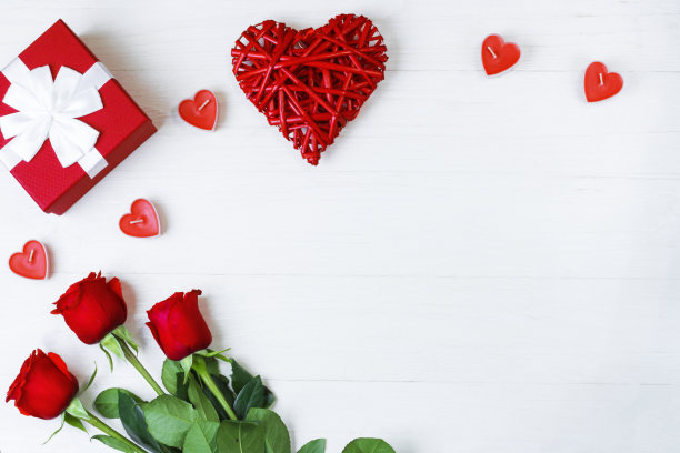 牛皮纸礼盒，上面有漂亮的白丝带和红玫瑰，心形的蜡烛，旁边是一个红色的牛皮纸心，在白色的木制背景上。副本的空间。前视图。图片下载
