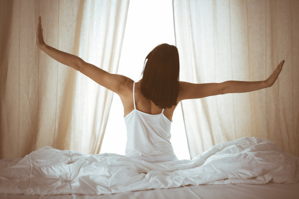 女人在床上伸开双手醒来后，太阳耀斑。深色头发的女人进入一个愉快和放松的一天后，睡了一个好觉和后视图。崭新的一天和快乐的周末图片下载