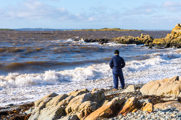 一个老人站在多岩石的海岸上，这是一个晴朗的冬天的早晨，涨潮了。图片下载