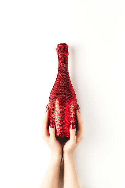 美丽的女性手与时尚的红色美甲持有香槟瓶在封面上闪耀着充满活力的红色亮片在白色的背景。模型为您的设计。副本的空间。平的。图片下载