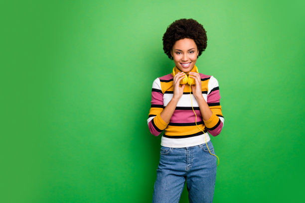 照片惊人的黑皮肤女士现代科技耳机耳朵听流行歌曲好心情穿休闲条纹毛衣牛仔裤孤立的绿色背景图片下载