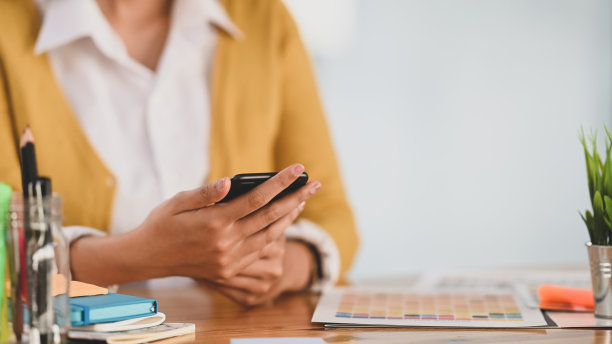 欢快的设计师坐在木制办公桌前，手里拿着黑色智能手机。图片下载