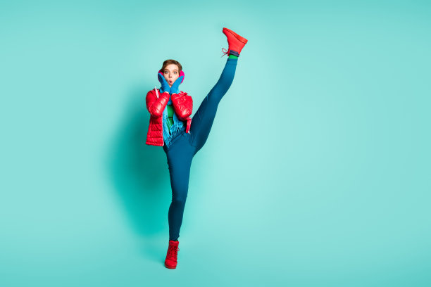 滑稽女士的全身照片，提高腿高，难以置信的灵活性，第一次健身训练后，穿红色大衣，手套，粉色耳套，裤子，鞋子，孤立的蓝绿色背景图片下载