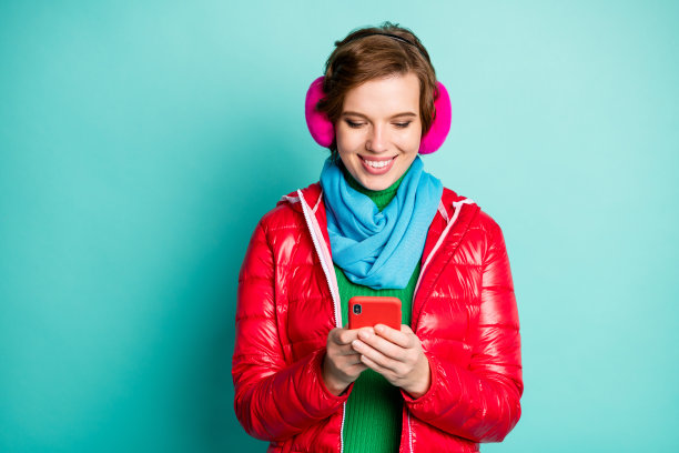 漂亮有趣的女士聊天的照片，电话，写信的朋友，在国外旅行穿休闲的红色外套围巾，粉红色的耳朵暖器，毛衣孤立的，蓝绿色的背景图片下载