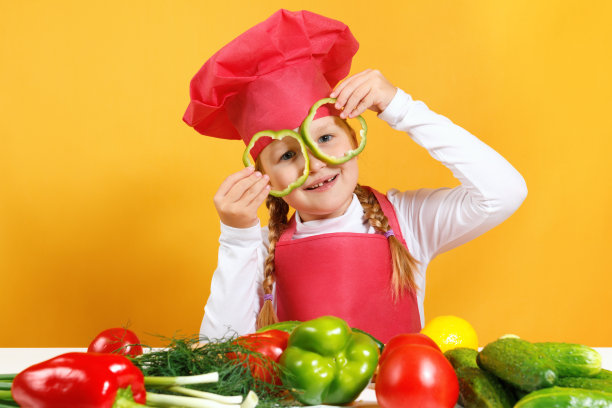 一个戴着厨师帽的快乐的孩子喜欢吃蔬菜，还把辣椒放在眼睛前，就像戴着眼镜一样。小女孩坐在一张以黄色墙壁为背景的桌子旁图片下载