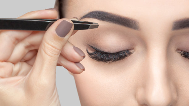 化妆师用镊子拔眉毛给一个有着棕色卷发和裸妆的女人。漂亮的浓眉合上。专业的化妆美容护肤。图片下载