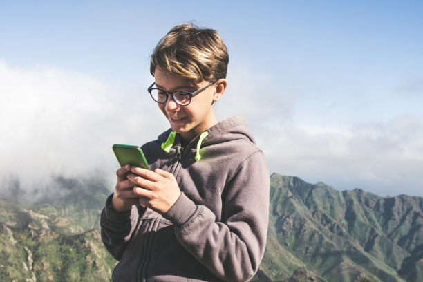 青少年正在用智能手机写短信，享受在公园户外度假的一天。在野外旅行中，微笑的男孩用手机分享视频。青春、科技的生活理念图片下载