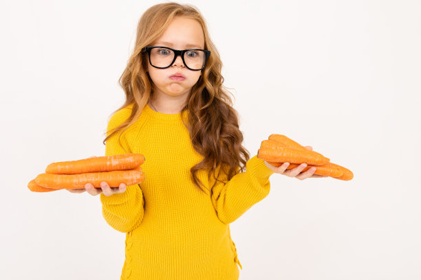 无聊的少女红头发，连帽衫和黄裤子手持胡萝卜孤立的白色背景图片下载