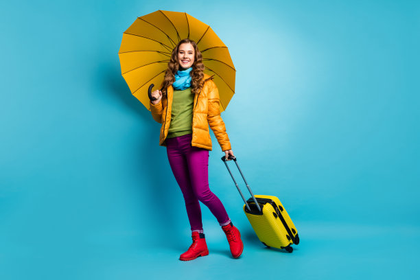 美丽的旅行者女士的全身照片撑伞滚动手提箱走机场登记穿黄色大衣蓝色围巾裤子靴子孤立的蓝色背景图片下载