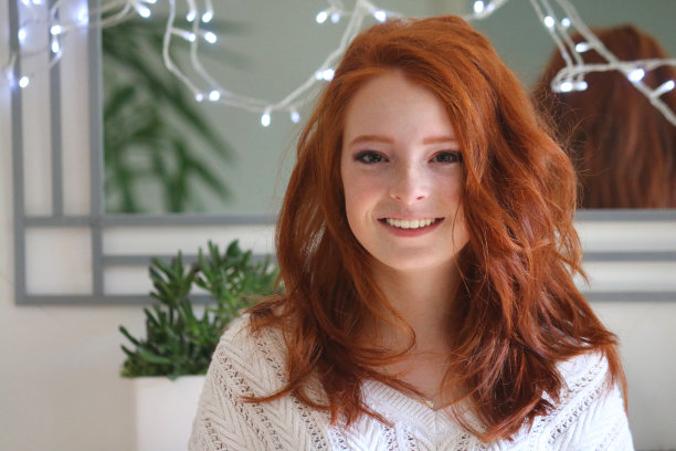 微笑的图像，红头发的少女14 / 15岁看着相机站在墙上的镜子由仙女灯照明，姜黄色的波浪发型，自然的化妆，苍白的皮肤和雀斑的stock照片图片下载