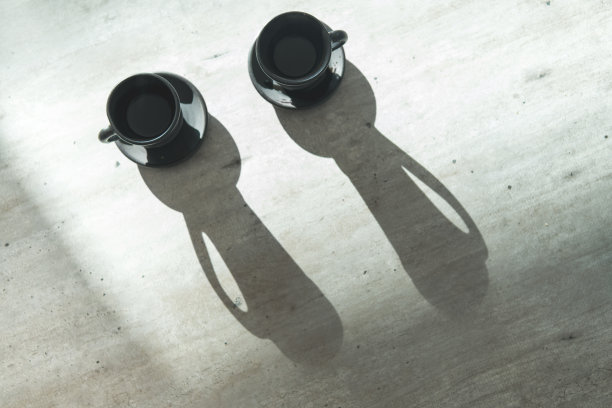 桌子上放着两个咖啡泥黑杯子，明亮的背光产生了长长的杯子阴影。现代厨房概念的情侣咖啡。图片下载