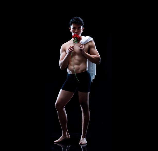 华裔年轻男脱衣舞娘身穿平角短裤，手持鲜花站在黑色背景前图片下载