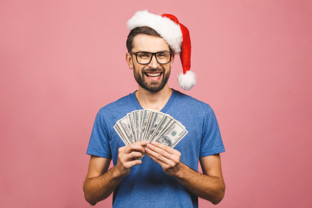 快乐的大胡子男人在休闲和圣诞帽拿着钱，看着孤立的粉红色背景的相机。图片下载