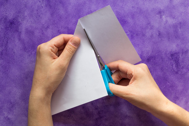 用剪刀剪开纸的边缘，用女人的手将纸的角折成方形，放在紫色的表面上图片下载