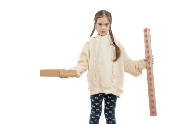 这个悲伤、不快乐的小女孩梳着辫子，穿着米色的连帽衫，左手拿着纸箱，右手拿着包装纸，在白色的背景下看着孤立的摄像机图片下载