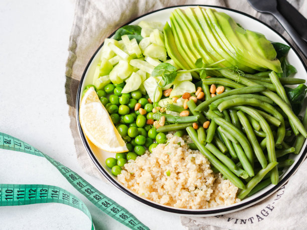 配有青豆、青豆、黄瓜、牛油果和松仁的藜麦碗，以及在灰色背景下测量身体体积的卷尺。图片下载