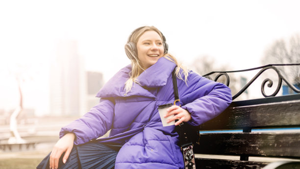 时尚美女肖像，身穿紫色外套，在街上戴着无线耳机听音乐，概念照片，手捧暖饮图片下载